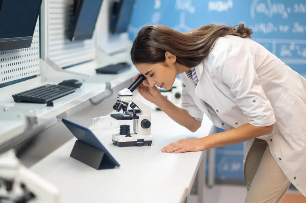 Zdravnica ali znanstvenica, ki proučuje vzorec preko mikroskopa, obkrožena z laboratorijsko opremo, prikazano v članku o ustnem zdravju.