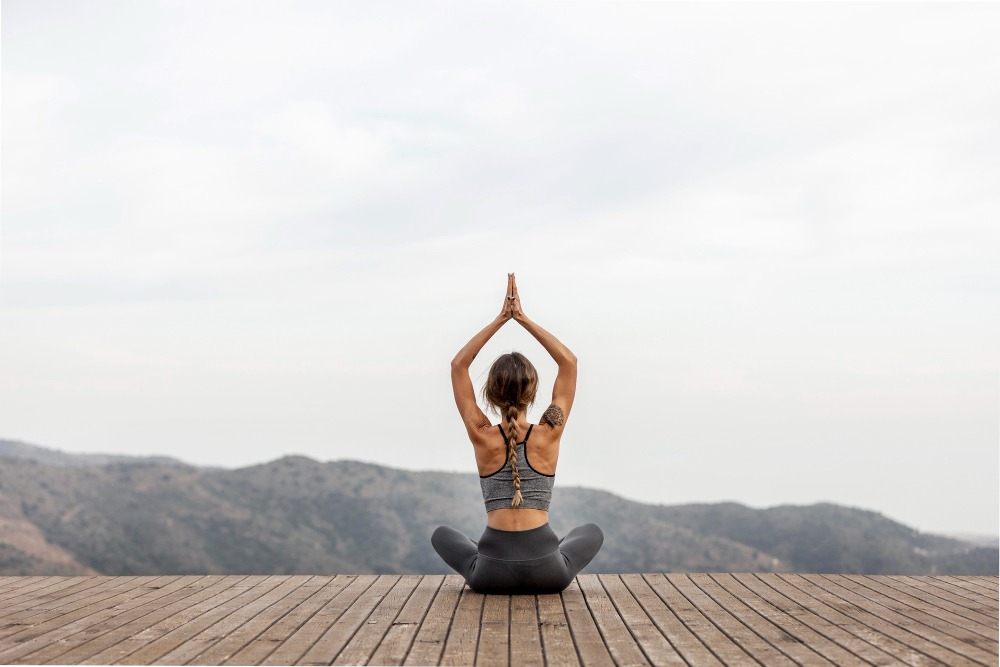 Ženska sedi v meditativni pozi na leseni palubi in zre v zeleno naravo pred seboj.