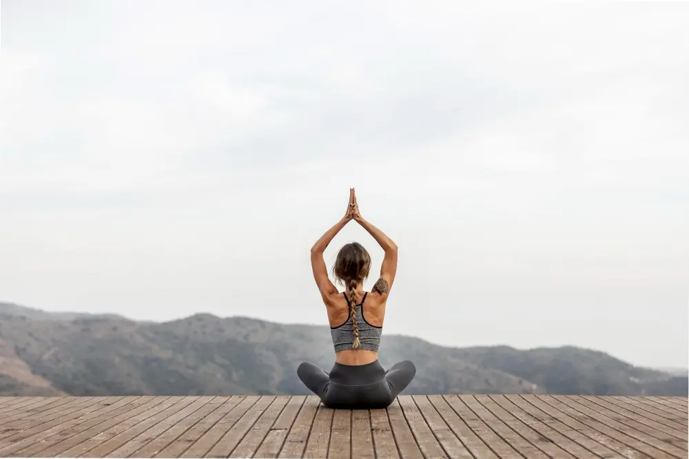 Ženska sedi v meditativni pozi na leseni palubi in zre v zeleno naravo pred seboj.